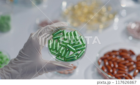 The researcher hand is holding a petri dish containing blue capsules. Samples of antibiotics, painkillers... Concept of researching medicinal products in the laboratory The researcher hand is holding a petri dish containing blue capsules. Samples of antibiotics, painkillers... Concept of researching medicinal products in the laboratory 117626867