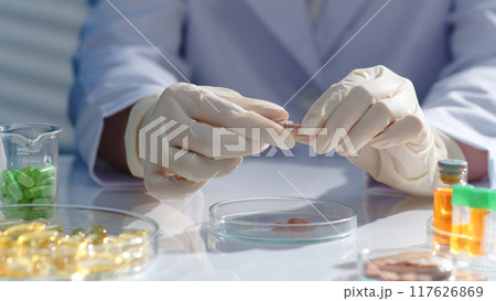 The researcher hands are holding a capsule and preparing to break it. Drug research scene, surrounded by lab glassware containing many different types of drugs. Scene for advertising 117626869