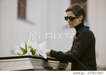 Senior widow in mourning clothes adjusting white lilies on black coffin next to church 117627007