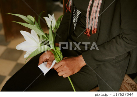 Unrecognizable woman dressed in black clothes sitting on wooden bench and holding white napkin with white lily during funeral Unrecognizable woman dressed in black clothes sitting on wooden bench and holding white napkin with white lily during funeral 117627044