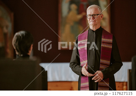 Silver-haired pastor dressed in black robe saying prayer during funeral 117627115