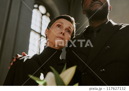 Senior Caucasian woman with tears in eyes holding madonna lily bouquet while leaning on sons shoulder in church 117627132