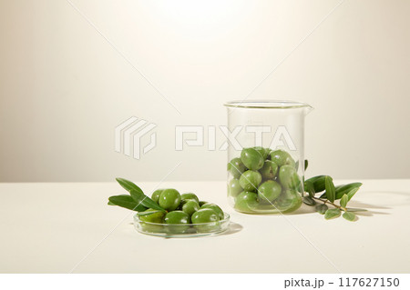 A front view of lab glassware adorned with fresh olives, arranged on a white table with a white background, advertises the olive ingredient. 117627150