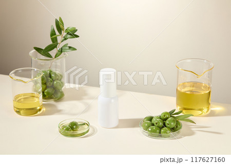 Cosmetic advertisement about olive ingredient, white cylindrical bottle placed in the middle of white table, surrounded by olive ingredients placed in glass laboratory equipment. Cosmetic advertisement about olive ingredient, white cylindrical bottle placed in the middle of white table, surrounded by olive ingredients placed in glass laboratory equipment. 117627160