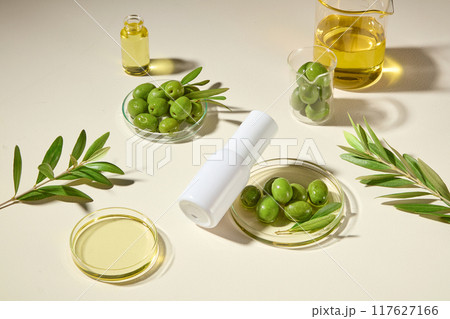 Top view, white cylindrical bottle is tilted onto a petri dish containing olives and essential oil, surrounded by olive branches and experimental glassware. Cosmetic natural theme. Top view, white cylindrical bottle is tilted onto a petri dish containing olives and essential oil, surrounded by olive branches and experimental glassware. Cosmetic natural theme. 117627166