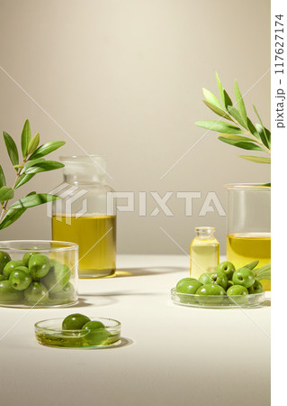 Olive fruit, olea Europaea oil are filled in transparent petri dishes, glass jars and beaker. Garnished with olive leaves to create a minimalist layout for advertising. 117627174