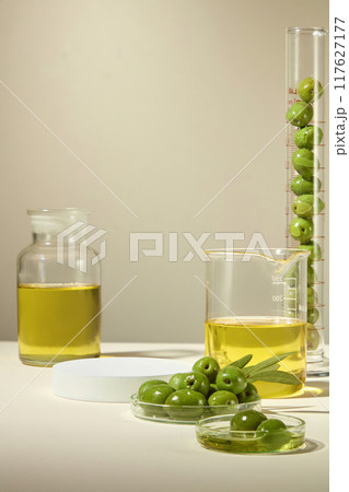 A tall graduated cylinder and petri dishes filled with green olives, beaker and glass jar filled with olive oil, empty white platform for products, ideal for cosmetics advertising. 117627177