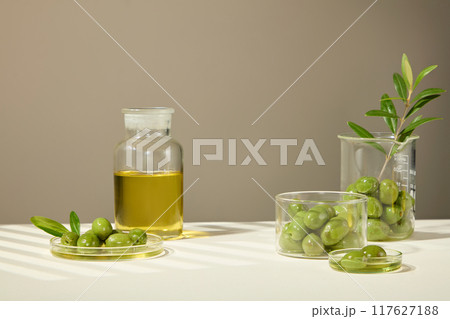 Olives and extract are placed in various containers such as beakers, petri dishes, and glass jars, with a designated space in the middle for the product. Cosmetics advertisement. 117627188