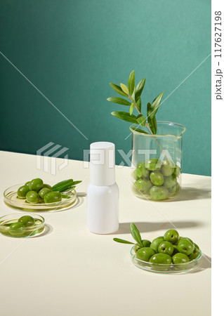 Simple cosmetic advertisement features a white cylindrical bottle, surrounded by petri dishes and beakers containing fresh green olives, olive leaves, and olive oil. 117627198