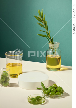 A transparent petri dish, beaker, and cork stopper jar contain fresh green olives, olive oil and branches, with a product display platform for cosmetic advertising. 117627199