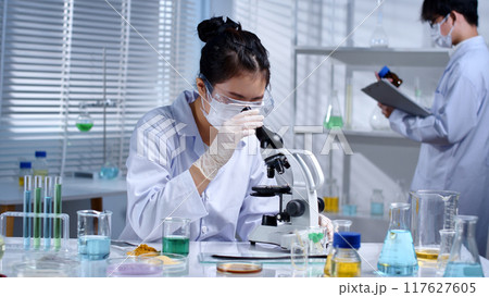 The scientist is looking at a microscope to study hair samples. Modern laboratory setting with chemicals and solutions to research hair care products, restore damaged hair 117627605