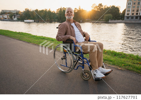 Happy senior man in wheelchair smiling enjoying freedom walking on road in city park outdoor. Old cheerful positive man in chair for people with disability. Disability disease rehabilitation 117627701