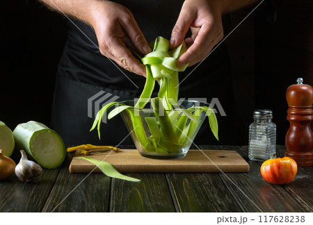 A chef fills a glass bowl with sliced zucchini before frying it with vegetables in the kitchen. Concept of making vegetable marrow for breakfast 117628238