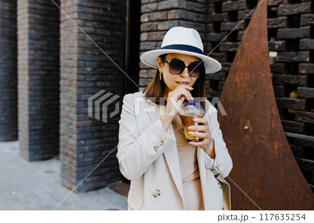 Portrait of successful business woman holding cup of cold drink in hand on her way to work on city street Portrait of successful business woman holding cup of cold drink in hand on her way to work on city street 117635254