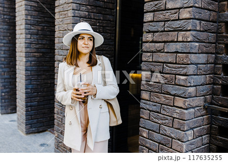 Portrait of successful business woman holding cup of cold drink in hand on her way to work on city street Portrait of successful business woman holding cup of cold drink in hand on her way to work on city street 117635255