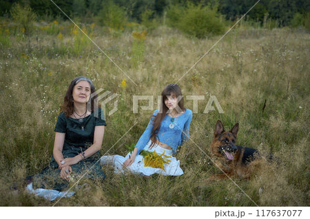 two woman in a whimsical aesthetic style dress with a german shepherd in a field in the summer at sunset two woman in a whimsical aesthetic style dress with a german shepherd in a field in the summer at sunset 117637077