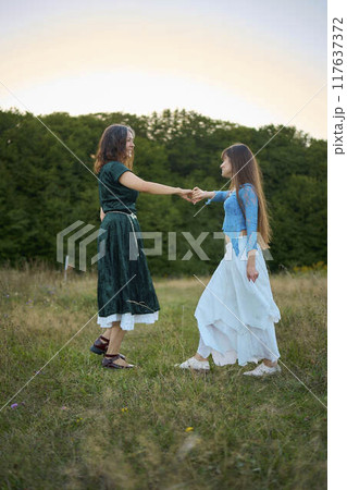 two woman in  whimsical aesthetic style dress  dance in a field in the summer at a sunset 117637372