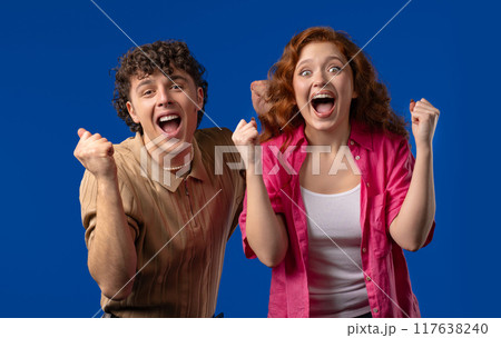 Pretty woman and man shows triumph yes yeah, hooray gesture of victory, achieved result, goals. Stylish couple glad, happy, surprised excited on blue background. 117638240