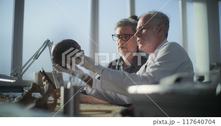 Paleoanthropologist holds prehistoric human skull, archaeologist uses tablet Paleoanthropologist holds prehistoric human skull, archaeologist uses tablet 117640704
