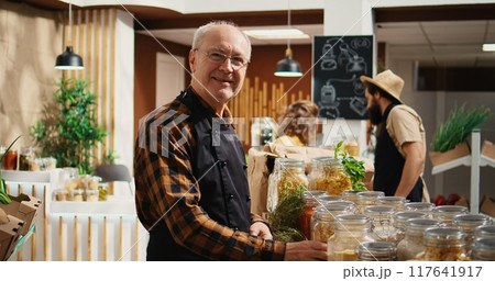 Portrait of elderly vendor working in zero waste organic food supermarket, adding pantry staples on shelves. Older employee restocks local grocery store with bulk products in biodegradable jars Portrait of elderly vendor working in zero waste organic food supermarket, adding pantry staples on shelves. Older employee restocks local grocery store with bulk products in biodegradable jars 117641917