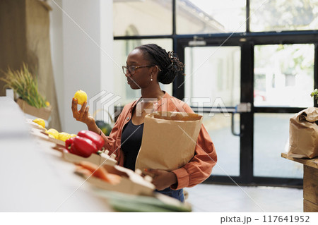 Young black woman holding fresh lemon and brown paper bag in eco friendly bio food shop. African american female customer examining freshly harvested produce in local convenience store. 117641952