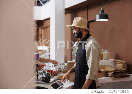 Middle eastern salesman using weighing machine to measure locally grown products for customer. At the counter, male vendor checking the weight and price of freshly harvested eggplant. Middle eastern salesman using weighing machine to measure locally grown products for customer. At the counter, male vendor checking the weight and price of freshly harvested eggplant. 117641986