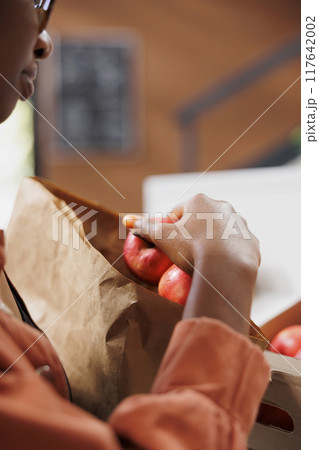 Close up of black person placing freshly harvested red apples in paper bag at grocery shop. Image showing african american female individual putting locally grown fruits in brown sack. Close up of black person placing freshly harvested red apples in paper bag at grocery shop. Image showing african american female individual putting locally grown fruits in brown sack. 117642002