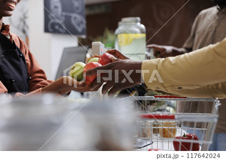 Close-up shot of hands of two african american people holding a bunch of apples over the checkout counter. Selective focus on female vendor receiving fruits from customer for weighing. Close-up shot of hands of two african american people holding a bunch of apples over the checkout counter. Selective focus on female vendor receiving fruits from customer for weighing. 117642024