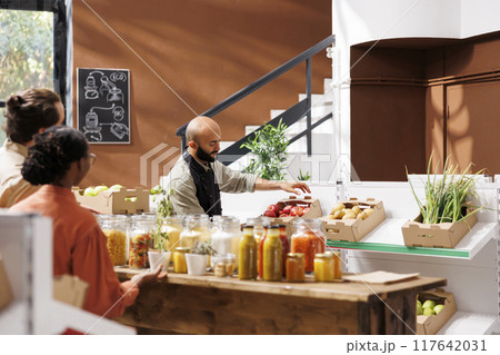 Middle Eastern storekeeper putting a box filled with red apples on shelf in eco friendly store. Male vendor arranges aisle of organic fresh products while two customers, male and female, look on. 117642031