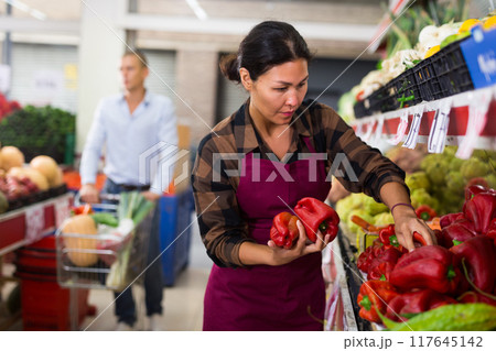 Woman in apron stacking red peppers in greengrocer 117645142