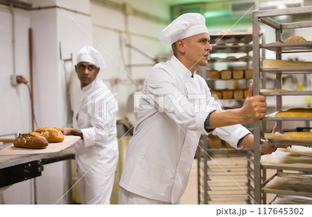 Male baker carrying rack trolley with bread Male baker carrying rack trolley with bread 117645302