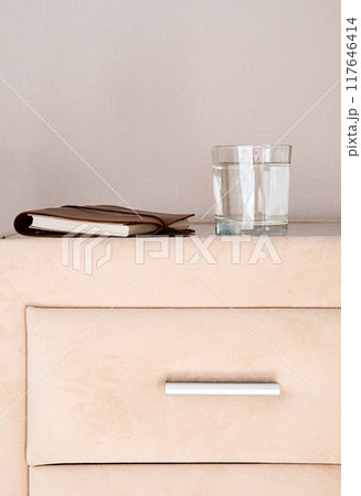 Leather bound notebook and glass of water on beige nightstand. 117646414