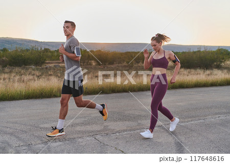 A handsome young couple running together during the early morning hours, with the mesmerizing sunrise casting a warm glow, symbolizing their shared love and vitality A handsome young couple running together during the early morning hours, with the mesmerizing sunrise casting a warm glow, symbolizing their shared love and vitality 117648616