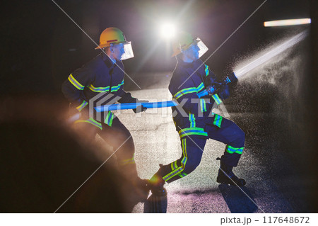Firefighters using a water hose to eliminate a fire hazard. Team of female and male firemen in dangerous rescue mission.  Firefighters using a water hose to eliminate a fire hazard. Team of female and male firemen in dangerous rescue mission.  117648672