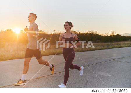 A handsome young couple running together during the early morning hours, with the mesmerizing sunrise casting a warm glow, symbolizing their shared love and vitality 117648682