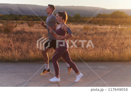 A handsome young couple running together during the early morning hours, with the mesmerizing sunrise casting a warm glow, symbolizing their shared love and vitality 117649058