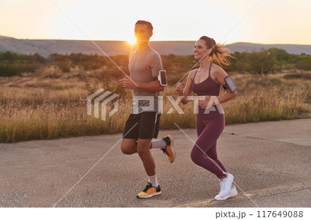 A handsome young couple running together during the early morning hours, with the mesmerizing sunrise casting a warm glow, symbolizing their shared love and vitality A handsome young couple running together during the early morning hours, with the mesmerizing sunrise casting a warm glow, symbolizing their shared love and vitality 117649088