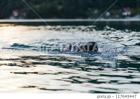 Triathlon athlete swimming on lake in sunrise wearing wetsuit 117649447