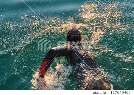Triathlon athlete swimming on lake in sunrise wearing wetsuit Triathlon athlete swimming on lake in sunrise wearing wetsuit 117649463