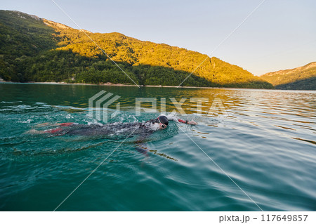 Triathlon athlete swimming on lake in sunrise wearing wetsuit 117649857