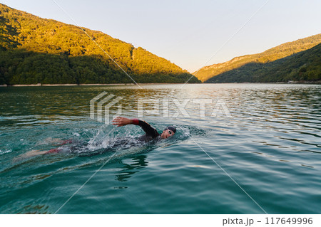 Triathlon athlete swimming on lake in sunrise wearing wetsuit 117649996