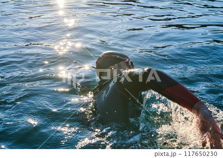 Triathlon athlete swimming on lake in sunrise wearing wetsuit Triathlon athlete swimming on lake in sunrise wearing wetsuit 117650230