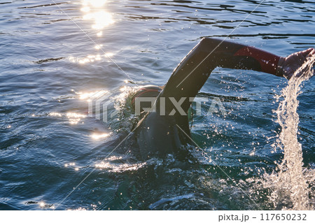 Triathlon athlete swimming on lake in sunrise wearing wetsuit Triathlon athlete swimming on lake in sunrise wearing wetsuit 117650232