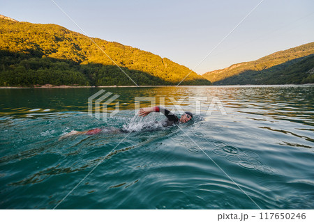 Triathlon athlete swimming on lake in sunrise wearing wetsuit Triathlon athlete swimming on lake in sunrise wearing wetsuit 117650246