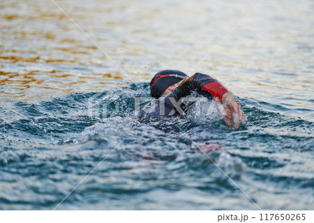 Triathlon athlete swimming on lake in sunrise wearing wetsuit Triathlon athlete swimming on lake in sunrise wearing wetsuit 117650265