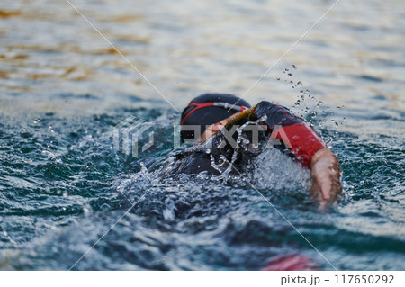 Triathlon athlete swimming on lake in sunrise wearing wetsuit Triathlon athlete swimming on lake in sunrise wearing wetsuit 117650292