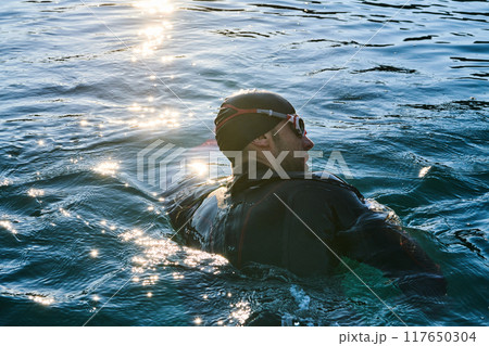 Triathlon athlete swimming on lake in sunrise wearing wetsuit 117650304