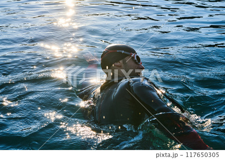 Triathlon athlete swimming on lake in sunrise wearing wetsuit 117650305