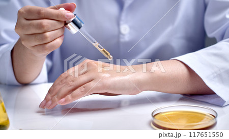 Close-up photo from the front of a laboratory scene, a scientist testing cosmetics on the back of his hand, next to a glass petri dish contains yellow liquid. Photo for advertising, space for text 117651550