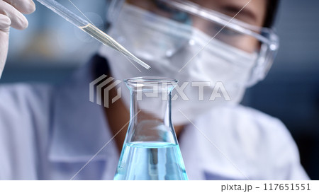 Macro front angle view of a scientist is dropping light blue liquid substance into an erlenmeyer flask. Concept of substance study experiment on blurred lab background, for promotional photo Macro front angle view of a scientist is dropping light blue liquid substance into an erlenmeyer flask. Concept of substance study experiment on blurred lab background, for promotional photo 117651551